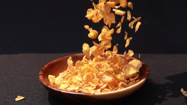 Pouring crispy yellow corn flakes into a wooden bowl on black background in Slow Motion