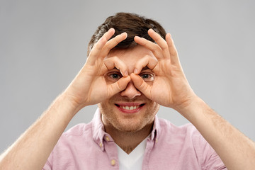 fun and people concept - smiling young man looking through finger glasses over grey background