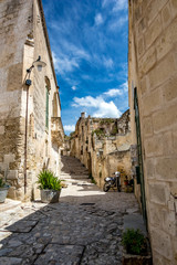 Fototapeta na wymiar Summer day scenery street view of the amazing ancient town of the Sassi with motorbike and white puffy clouds moving on the Italian blue sky. Matera, Basilicata, Italy