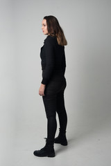 full length portrait of a brunette girl wearing  modern black jacket and pants, standing pose with back to the camera on grey studio background.