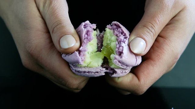 closeup human hand break in half violet french dessert macaroon with green creamy filling on black mirror background