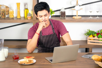 Asian young man using laptop for online working at home with toast  strawberry jam and milk on wooden table for breakfast in modern living room.