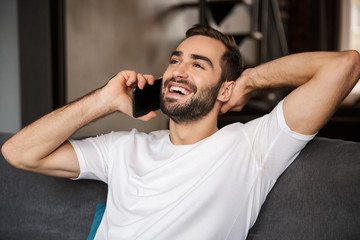 Photo of joyful man talking on smartphone while sitting on sofa in living room