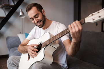 Photo of positive man playing acoustic guitar while sitting on sofa in apartment