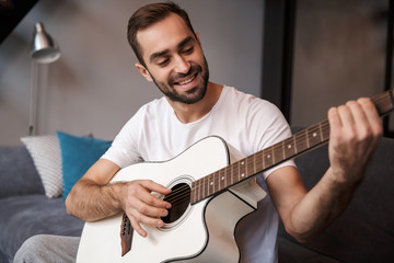 Photo of attractive man playing acoustic guitar while sitting on sofa in apartment