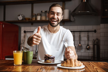 Portrait of joyful man 30s eating while having breakfast in stylish kitchen at home