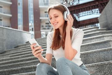 Young girl in headphones sitting on stairs in the city with smartphone listening music closed eyes...