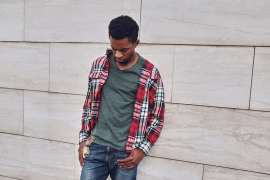 Stylish african man wearing red plaid shirt looking down, young guy posing on city street, gray brick wall background