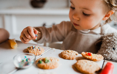 A small toddler girl sitting at the table, decorating cakes at home.