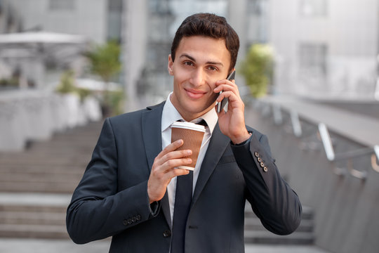 Morning refreshment. Businessman standing on the city street ddrinking coffee talking on smartphone smiling happy close-up