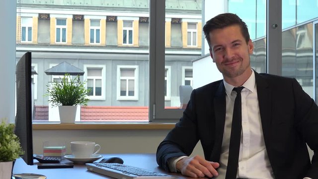 An office worker in a suit sits at a desk in front of a computer and shows a thumb up to the camera