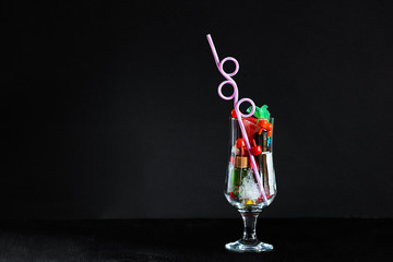 Coctail glass with plastic straw full of plastic and battery waste as imitation of real drink on horozontal black background with copy space. Ecology concept of getting pollution to food and drinks