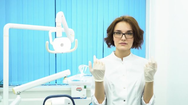 Young woman doctor wearing glasses and a white coat, putting on white medical glovesin dental clinic cabinet. A strict female dentist puts on gloves and looks into the camera, getting ready.