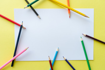 A rainbow of colored pencils are scattered randomly on a white sheet of paper that is lying on a yellow background. Top view