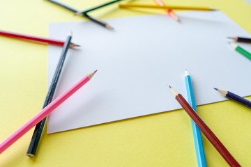 A rainbow of colored pencils are scattered randomly on a white sheet of paper that is lying on a yellow background.