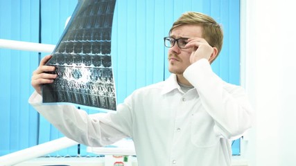 Male doctor radiologist looking at x-ray, touching his glasses in medical cabinet, medicine concept. Young practitioner man standing in hospital cabinet and looking at radiograph.