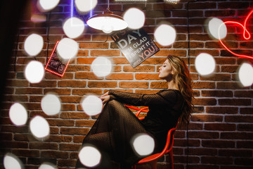 Resplendent girl in a black dress sitting on a chair near a brick wall