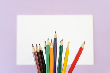 Close-up of a rainbow of colored pencils for child art above the white piece of paper on purple background. Top view