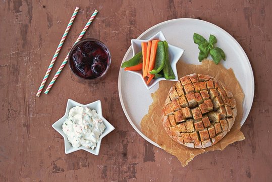 Party set on a brown concrete background: baked party bread with herbs and olive oil, a dip of cream cheese with vegetables, sliced raw carrots and green sweet pepper, grape juice with ice, straw.
