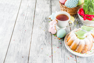 Easter table setting concept, festive table with decoration of young grass, cake, pastel colored eggs, homemade cookies in shape of eggs, bunny rabbits. On a wooden background, copy space