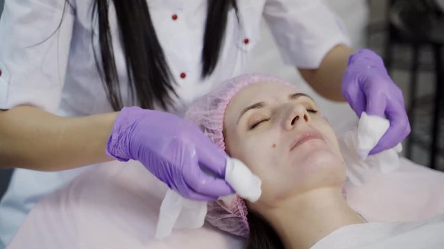 A beautician in gloves holds sanitary napkins in her both hands and cleans the skin of the client's face with it before the procedure. Close-up.