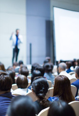 "Speaker on Stage at Conference Meeting Event. Presenter at Business Seminar Photo. Audience Watching a Manager Presentation. Blurred Image of Lecturer Presenting To Audience During Speech. "