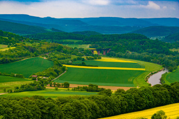 Naklejka premium Nice panoramic view of the Werra Valley landscape. The river Werra flows through cultivated canola fields and a railway bridge, surrounded by woodland and a low mountain range in the background.