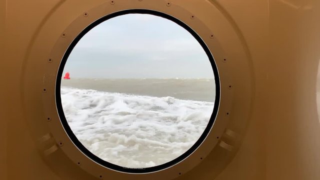 View through a porthole on a stormy sea with waves hitting the ship sailing past a red buoy at the Wadden Sea between Lauwersoog and Schiermmonnikoog
