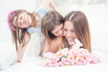 Obraz na płótnie Canvas Happy mother's day concept. Mom with twocute young daughters twins on the bed in the bedroom and a bouquet of flowers tulips