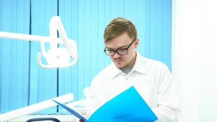 Portrait of doctor reading a report document at his office, shaking head, bad news and medicine concept. Young, serious and sad doctor or dentist who is looking at the opened blue folder indoors.