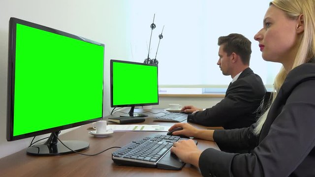 Two office workers, man and woman, work on computers with green screens