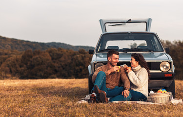 Beautiful young couple enjoying picnic time on the sunset. They drinking tea and sitting in a meadow leaning against a old fashioned car.