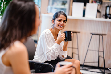 A beautiful Indian woman is chatting with a colleague inside a cafe. She is listening to a discussion while drinking hot coffee.