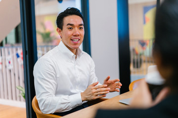 A youthful Asian Chinese expert is giving an introduction before his partners in the workplace. He is wearing a white polo shirt and a really nice hair do.