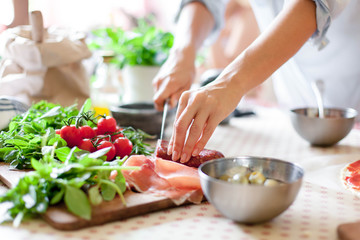 Woman is cooking in home kitchen. Female hands cut salami, vegetables, greens, tomatoes on table on wooden boards. Ingredients for preparing italian or french food. Lifestyle moment. - obrazy, fototapety, plakaty