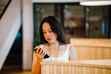 A closeup shot of a popular and gregarious, casual wearing Eurasian woman looking seriously at her mobile phone while standing beside a modern styled room divider.
