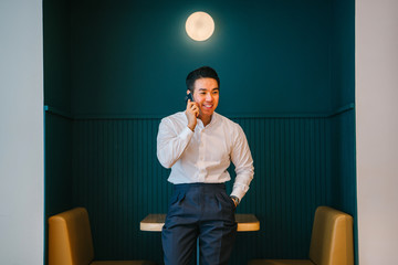 Portrait of a young, handsome and fit looking Chinese Asian man professionally dressed leaning against a table during the day. He is speaking on his smartphone and smiling.