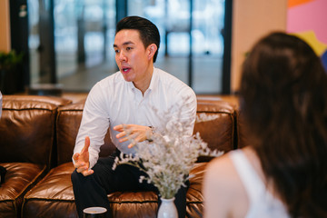 A youthful Asian Chinese expert is giving an introduction before his partners in the workplace. He is wearing a white polo shirt and dark jeans.