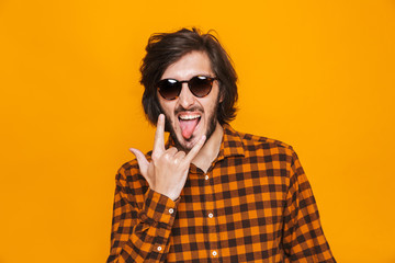 Portrait of european guy 20s wearing plaid shirt and sunglasses smiling and showing rock sign while standing isolated