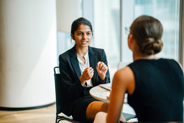 An exquisite Indian woman sitting by an accomplice inside a cafeteria. She is telling a joke with a...