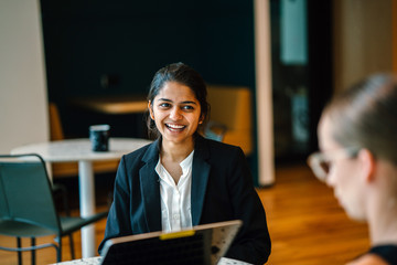 A lovely Indian lady sitting next to a partner inside a cafeteria. She is telling a joke with a collaborator.