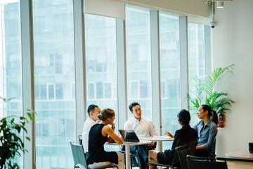 A diverse team sits around a table in a meeting room to have a business meeting to discuss plans. The group is international with Asian and white team members and they are all professionally dressed.