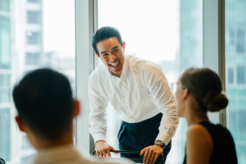 A gorgeous, certain and fit Asian Chinese man seats a gathering with his group amid the day in the workplace. He is expertly wearing a shirt and pants and is motioning as he talks.