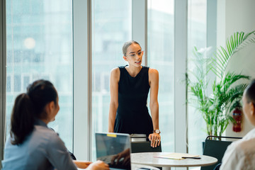 A young, professional Caucasian woman is formally presenting in front of her colleagues in a meeting room. She is professionally speaking wearing eyeglasses and a black top with white pants.