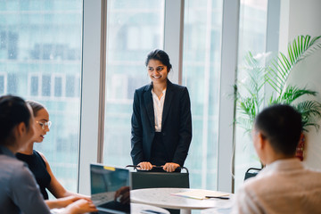 An Asian Indian businesswoman is having a conversation with her colleagues in a conference room. She's smiling elegantly.  She's wearing a black blazer and white blouse with a black skirt.