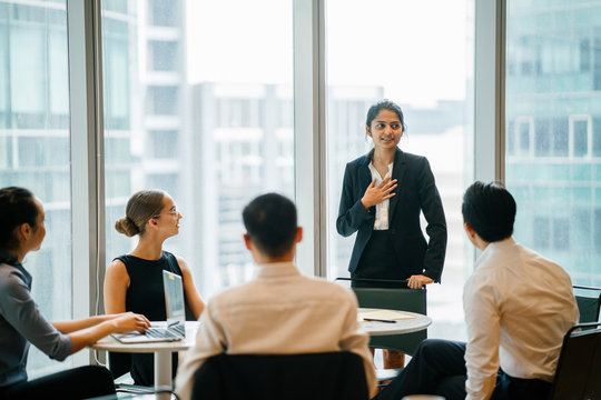 An Asian Indian businesswoman is ingeniously explaining with her diverse team in a conference room. She's wearing a black blazer and white blouse with a black skirt.