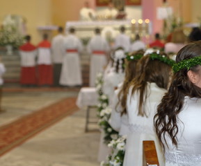 Church interior during First Holy Communion celebrating, girls in white dresses, with green wreath on heads sit on decorated with white flowers benches, some in soft focus, main altar, priests, candle