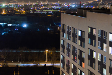 Fototapeta na wymiar High-rise modern building in the evening window lights.Casual urban city life. Traffic on the road.From tower rooftop, industrial region with working factory and container crains