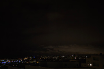 Fototapeta premium Night city skyline. Top aerial panoramic view of modern city from tower rooftop. Road junction traffic. Heavy clouds on the dark sky