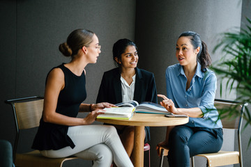 Portrait of a diverse group of three professionally dressed young woman (Chinese, Indian and Caucasian) having a casual discussion around a table in a meeting room during the day.